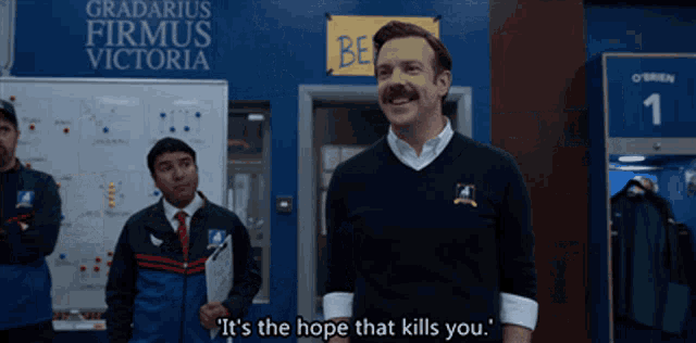 a man with a mustache is standing in front of a sign that says  it 's the hope that kills you 